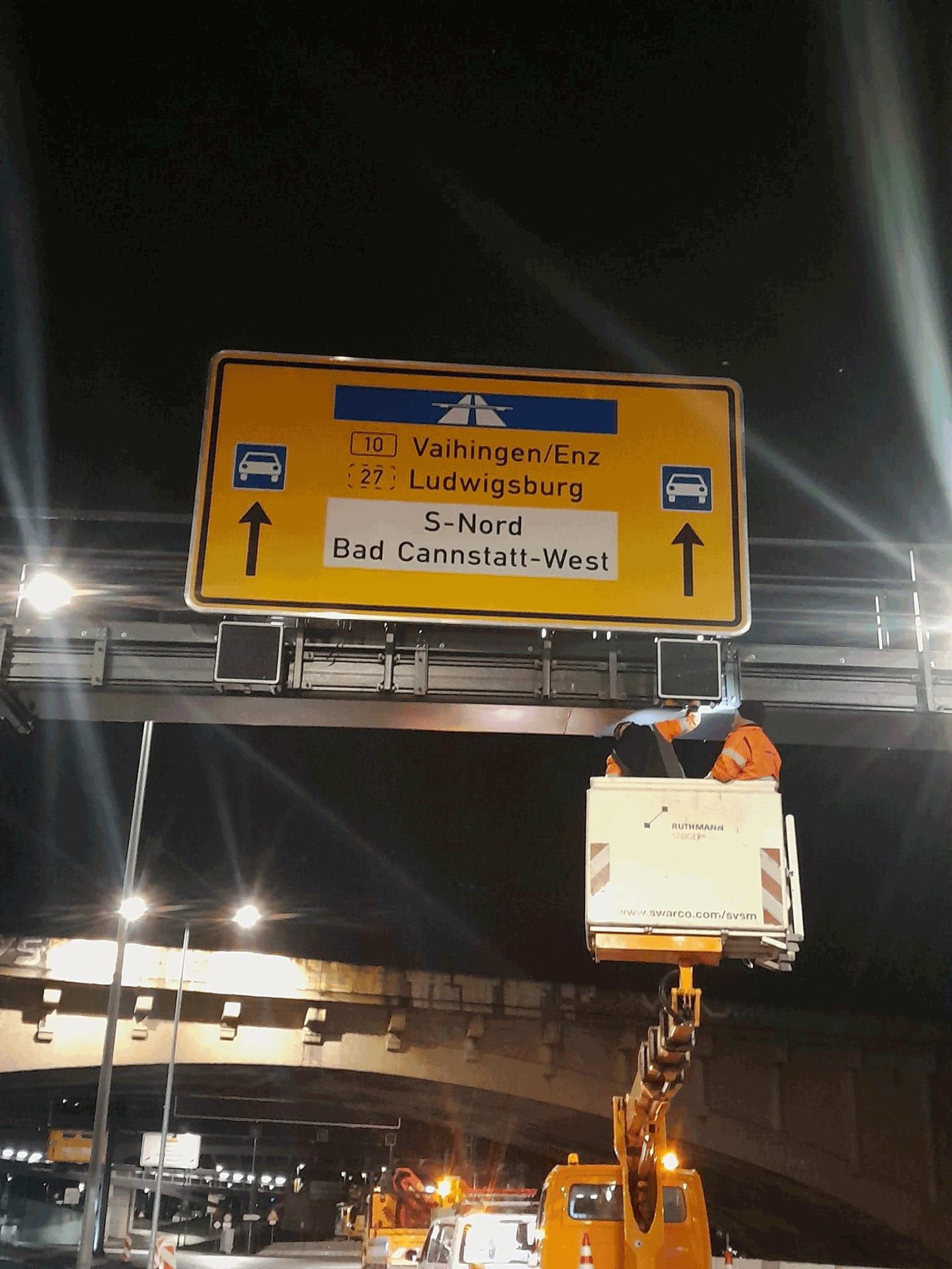 Nächtliche Montage eines Wegweisers an einer Verkehrszeichenbrücke mit Hubsteiger
