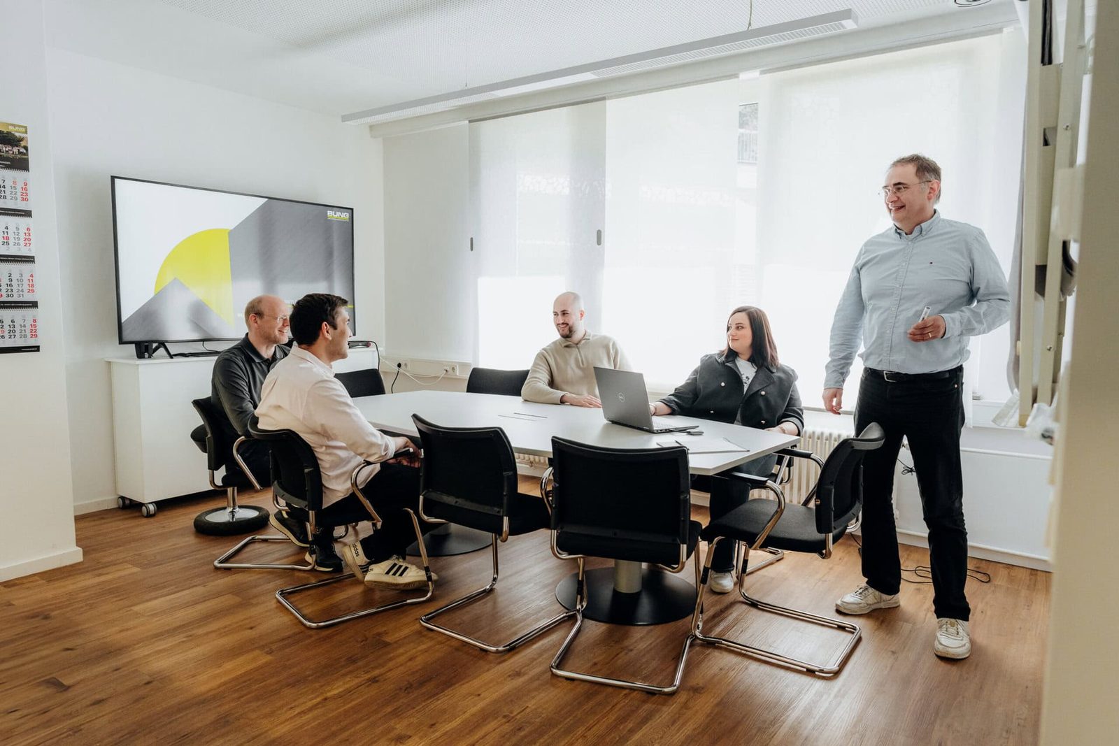 Besprechung an einem Konferenztisch, eine Person erläutert etwas im Stehen